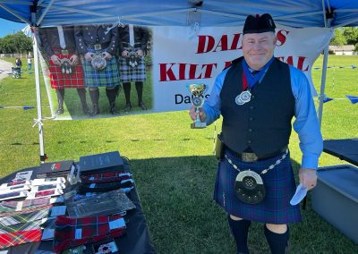 Kilted staff member holding a trophy at the Dallas Kilt Rental tent at a Texas Highland festival